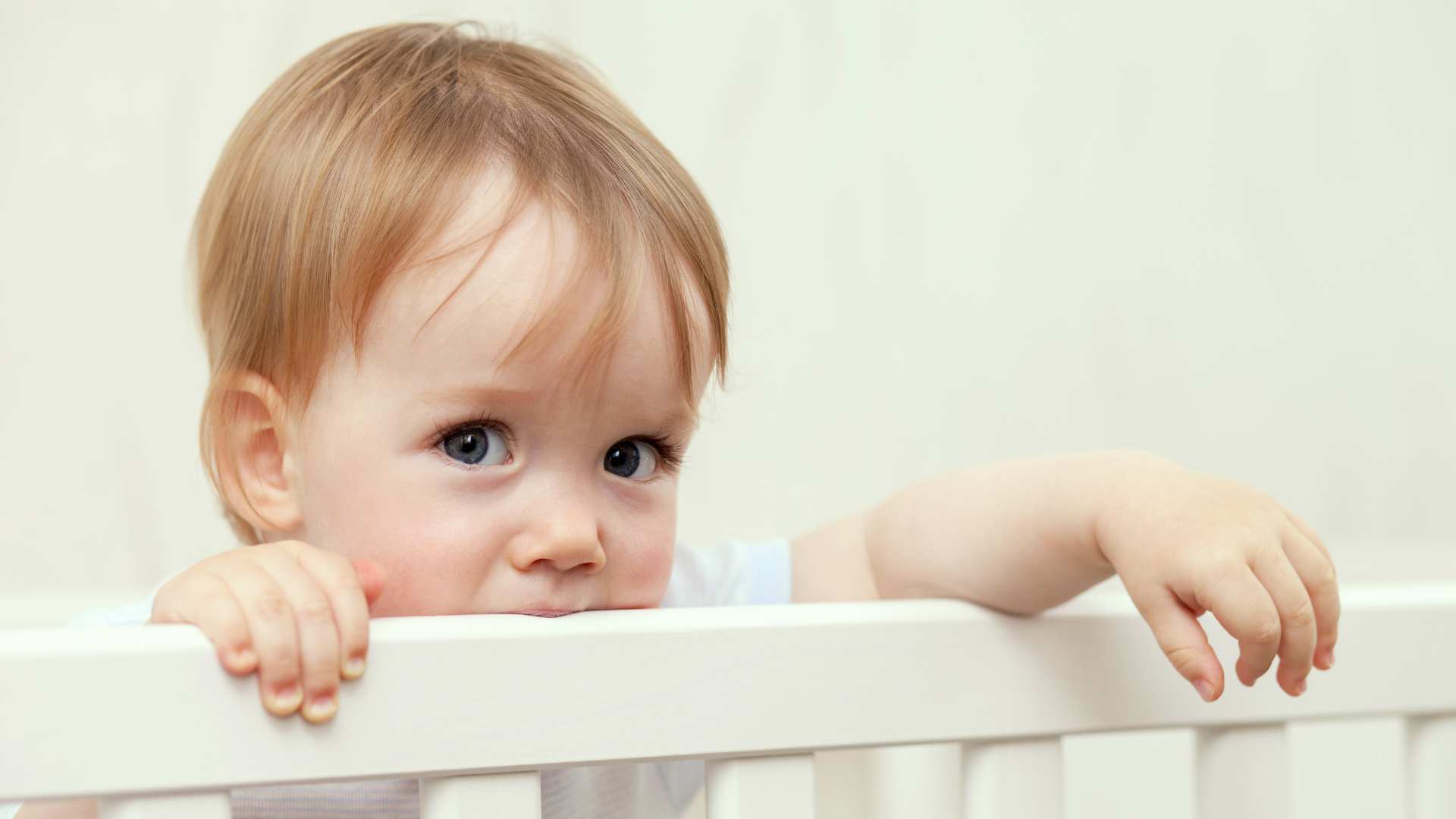 A baby standing in a crib in a calm indoor setting. The image is used to illustrate information about Signs of Autism at 12 Months