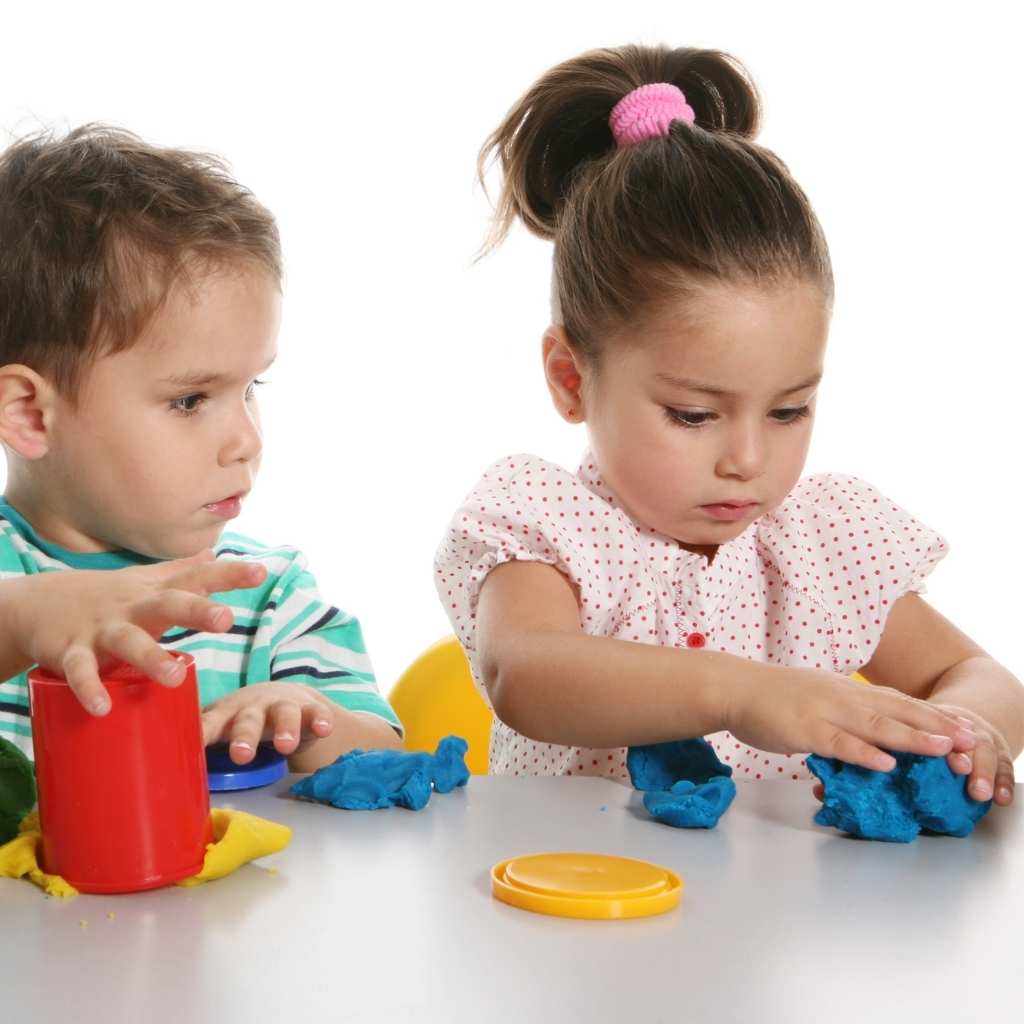 A child focused on shaping playdough while another child sits nearby, showing limited interest in peers, often seen in signs of autism at age 3.