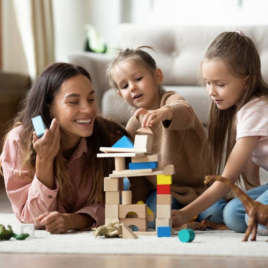 A toddler lines up toys next to an adult and two children playing with blocks, showing how parents can guide play by staying close, observing, and adding ideas to play