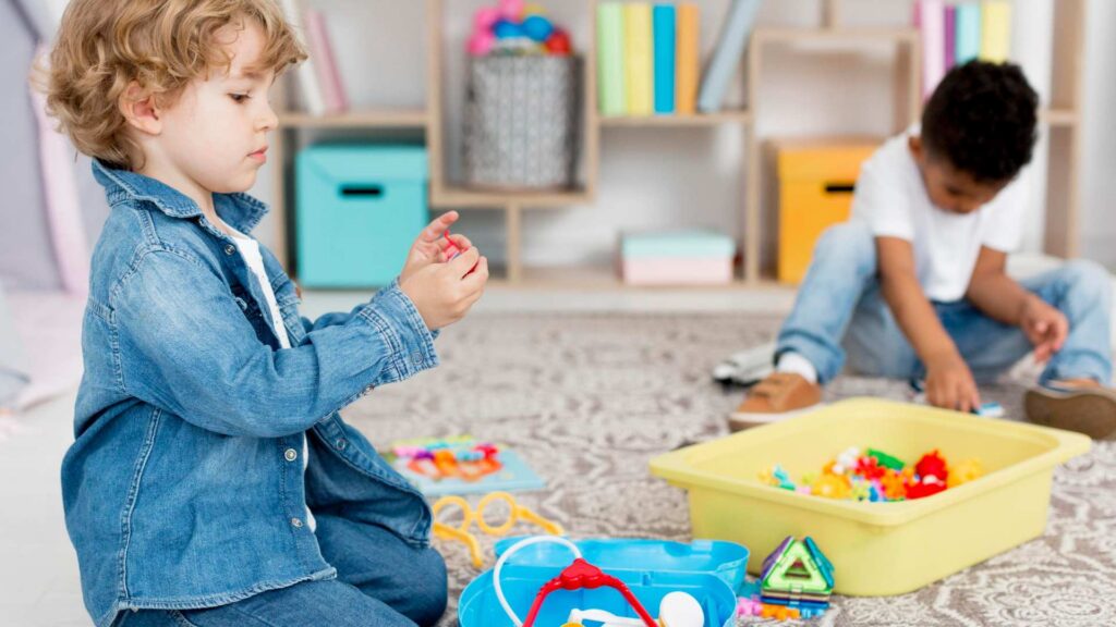 Two young children play separately with toys in the same room, illustrating signs of autism at age 3, such as not showing interest in peers.
