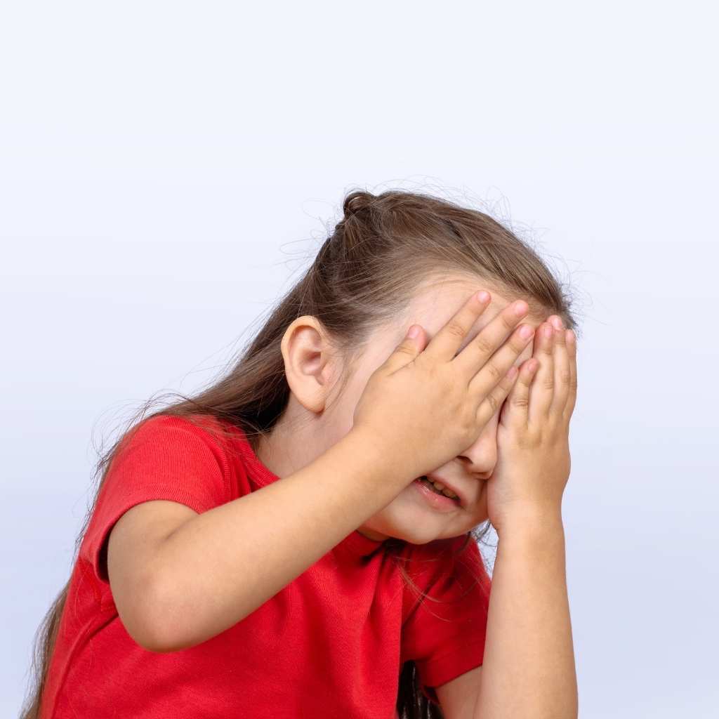 A young child in a red shirt covering their face with both hands, showing a moment related to eye contact in autism.