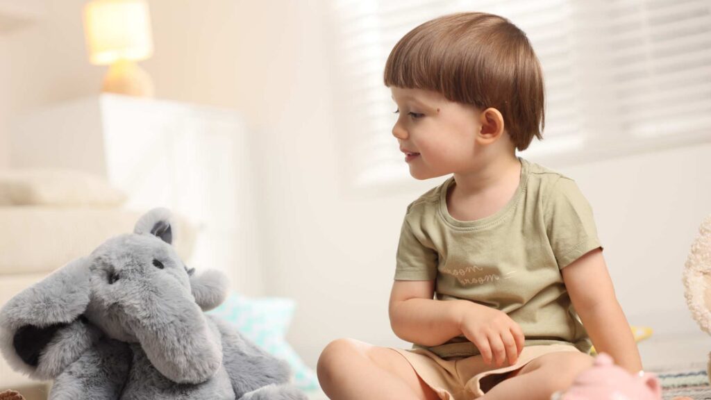 Toddler sitting on the floor looking at a stuffed animal, showing early communication behaviors.