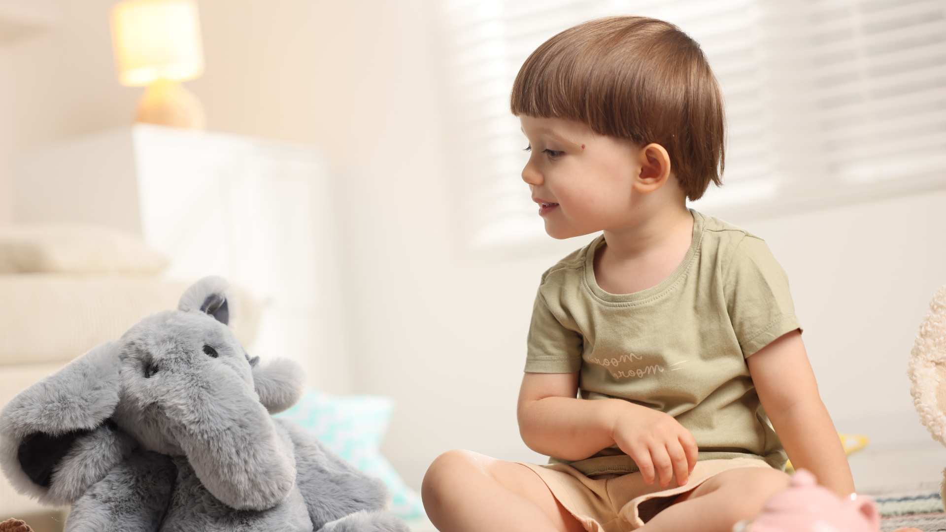 Toddler sitting on the floor looking at a stuffed animal, showing early communication behaviors.