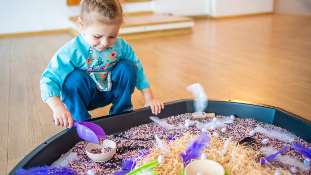 A toddler scooping materials from a sensory play bin filled with colorful textures